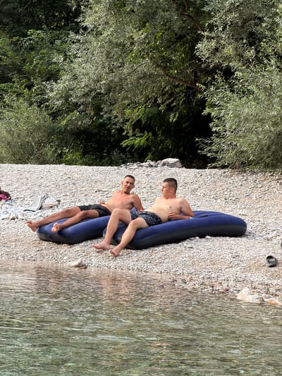 Two young men are on an inflatable raft on a rocky beach next to a clear river. The man closer to the camera is lying on his back with his head resting on the raft, wearing black swim trunks. The other man is sitting up on the raft, looking at the camera, and is wearing patterned swim trunks. He has his arm around the other man's shoulder. Lush green trees are visible behind them, suggesting a natural, outdoor setting. The water appears clear and shallow near the shore. The sun is shining, indicating a bright, warm day, likely during daylight hours. The scene conveys a sense of relaxation and enjoyment on a summer day. There is no visible text in the image.