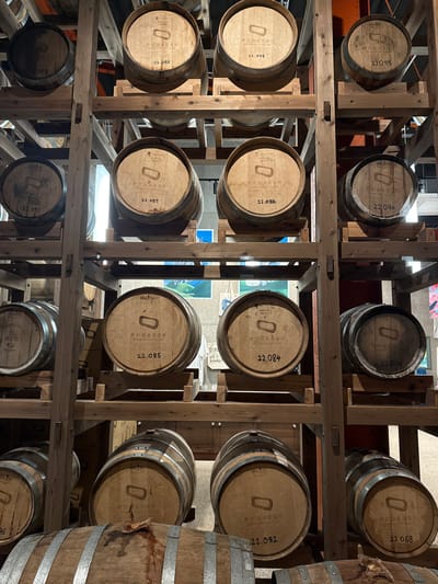 The image depicts an indoor scene within the Nozawa Onsen Distillery in Nozawaonsen, Japan, showcasing a multi-tiered wooden racking system holding numerous cylindrical wooden barrels. Each barrel face prominently displays a stenciled black emblem resembling a carabiner or elongated loop, above which Japanese text identifies "野沢温泉蒸留所" (Nozawa Onsen Distillery). Below the emblem, a four-digit identification number, such as "22.087" or "22.084," is visible on most barrels. The barrels vary in color from light to medium brown, with some exhibiting darker staining or discoloration, particularly on the barrel positioned at the bottom center, suggesting liquid seepage. The racking system is constructed from light-colored wooden planks and vertical support beams, providing horizontal cradles for each barrel. The background, out of focus, shows ambient lighting and what appear to be posters or graphic displays on a distant wall. No individuals are present, and no actions are occurring within the static scene.