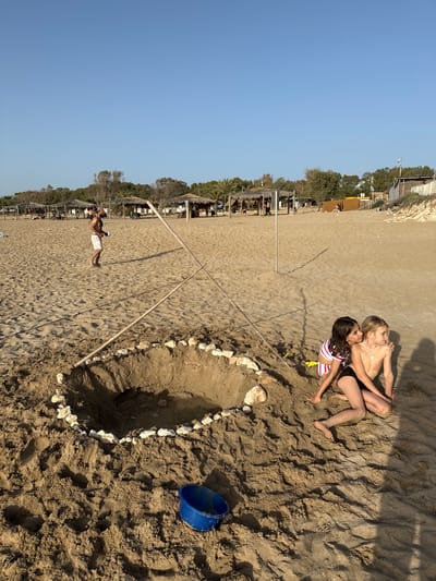 On a sandy beach in Nahariah, Israel, two girls are lying down together near a circle made of stones, next to a blue bucket. A man in white shorts is lying further down the beach near beach umbrellas. Two wooden poles form an X in the sand. In the background, there are trees and other structures. The sky is clear and blue.