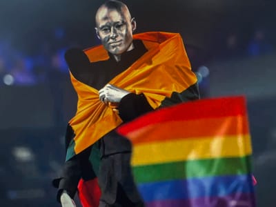 A person with their face and body painted silver is draped in an orange fabric. They are holding a rainbow flag. The background is dark and appears to be an indoor setting with blurred lights. The image is a close-up shot focusing on the individual and the flag. There is no information available regarding the specific location in the city of Ellwangen, Germany.