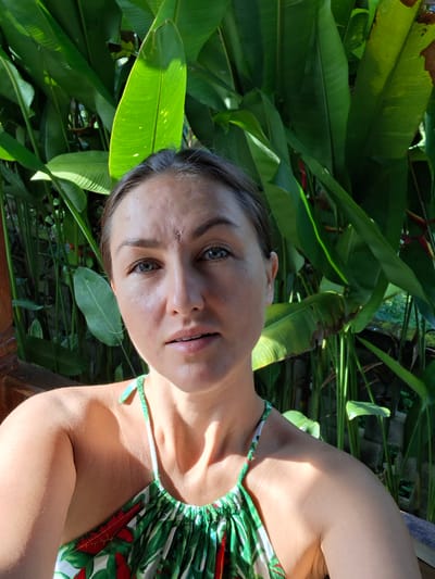 The image is a selfie of a woman with fair skin and light-colored eyes, possibly gray or blue-green. She has dark hair pulled back from her face. A small scar is visible on her forehead near her hairline. She is wearing a green and white floral patterned top with thin straps. The background is a lush green environment, filled with large tropical leaves and some red flowers, typical of tropical foliage. The image was taken in the city of Lodtunduh, Indonesia.
