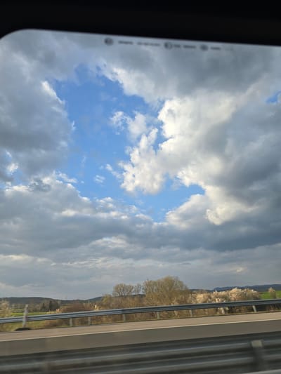 The image displays an exterior landscape observed from within a vehicle, indicated by a dark frame visible at the top. The sky is dominated by a mix of dark grey and white cumulus clouds, interspersed with clear patches of blue sky, suggesting a dynamic weather condition. In the foreground, a multi-tiered metal guardrail, consistent with highway infrastructure, extends horizontally across the lower portion of the frame, with a segment of an asphalt road and a white lane marking visible beneath it. The midground features a rural landscape characterized by various deciduous trees and bushes. Some trees exhibit nascent light-green foliage, while others are covered in prominent white blossoms, indicative of springtime. Green fields are present throughout this section. The background consists of undulating hills or distant mountains under the prevailing cloudy sky. No individuals or specific actions are discernible within the frame. This scene is located in the city of Yazdach, Bulgaria.