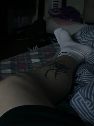This is a low-light, close-up, first-person perspective image of a person lying in bed. The focus is on the person's leg, which has a black spider tattoo on the calf and is covered by a white sock. The leg rests on bedding with a striped pattern in blue and white. In the background, dark shadows obscure much of the room, but hints of pillows, blankets, and what appears to be electronic equipment are visible. The image was taken inside a bedroom. There is no information in the image that allows for identification of the location in Muravlenko, Russia.