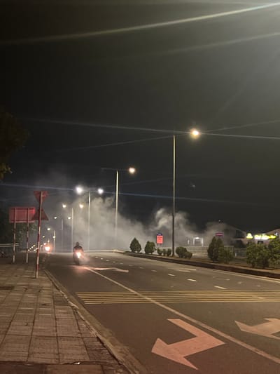 The image depicts a nighttime scene in Hoi An, Vietnam, featuring a street with various lights and a significant amount of smoke or fog. The perspective is from the side of the road, looking towards the street and sidewalk.

The primary subjects are the elements of the urban environment. Streetlights with bright bulbs are visible, casting light into the smoky atmosphere. The road itself has clear lane markings, including yellow lines and large white arrow indicators pointing in a direction that suggests traffic flow. A sidewalk with tiled paving runs alongside the road. In the background, there are blurred buildings and trees, illuminated by the streetlights and possibly lights from the structures.

A noticeable activity is the presence of smoke or fog that drifts across the road and sidewalk, creating an ethereal and somewhat obscured view of the scene. There is a person visible on the right side of the image, near the edge of the road, illuminated by a bright light source that appears to be on their person or very close to them. The light is fiery, suggesting a possible event involving flames or a bright flare.

Notable details include the time of day, clearly nighttime due to the darkness and artificial lighting. The smoke or fog suggests potentially humid or misty conditions, or it could be a result of an event. The location cues, such as the architecture and road markings, are consistent with a Vietnamese urban setting, specifically Hoi An. The overall mood is somewhat mysterious and dramatic due to the interplay of light and smoke.

There is no clearly legible text visible in the image. The focus is on the visual elements of the street, lighting, and the smoky atmospheric conditions.
