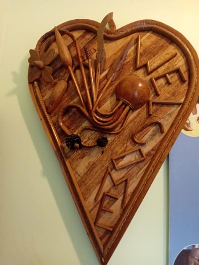 A close-up, eye-level shot captures a heart-shaped wooden carving displayed on a pale green wall. The carving is intricately detailed with various elements. In the upper left quadrant, a stylized wooden flower is visible, accompanied by what appear to be dried seed pods or reeds. A smooth, round, brown, bulbous object, possibly a gourd or polished wood, is positioned in the center, with curved wooden elements extending from it, resembling tentacles or vines. Towards the right side of the carving, the letters "L," "I," "F," and "E" are carved in a rustic, angular style. Below this, further down the heart shape, the letters "A," "N," and "D" are also carved. The carving features a textured, woven wooden border around its perimeter. Two small, dark, cone-shaped objects are positioned in the lower center of the carving. A poster or picture with a blue background and a partially visible circular element is seen to the right of the carving, suggesting it is displayed in a home or public space in Douala V, Cameroon. The lighting casts a shadow of the carving onto the wall to the left.