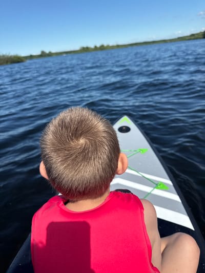 A low-angle, close-up shot shows a young boy from the shoulders up, facing away from the camera, looking out at a body of water. He is wearing a bright red life vest. His hair is light brown and styled in a swirl pattern at the crown. The boy is positioned on what appears to be a paddleboard or kayak, the front of which is visible in the lower right quadrant of the image. The board is white with gray stripes and green bungee cords. The water is a deep blue with visible ripples. In the distance, a tree-lined shore is visible under a clear blue sky. The location is the city of Ozerki, Russia.