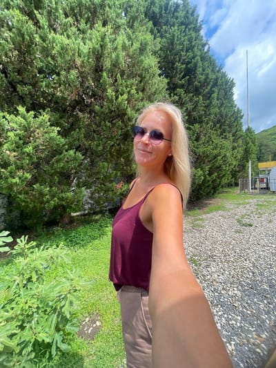 A blonde woman in sunglasses and a maroon tank top stands outdoors, taking a selfie. She is positioned in the foreground, with lush green trees and a cloudy sky as the backdrop. The ground is covered with small, light-colored pebbles. In the background, a fence and some structures are visible, along with a tall pole. The scene appears to be in a natural or semi-rural setting, possibly in or near Agoy, Russia.