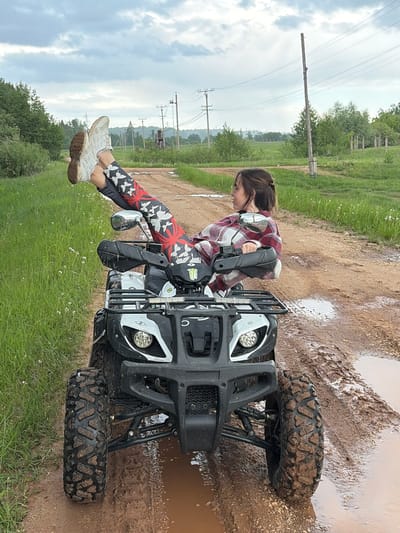 The image depicts a young woman seated on an all-terrain vehicle (ATV) in a muddy outdoor setting. The ATV is positioned on a dirt path with puddles, and the front tires are visibly coated in mud. The surrounding environment includes grass, trees, and a power line pole in the background. The woman is wearing a red and white plaid shirt, patterned leggings, and sneakers. She is holding the handlebars of the ATV, suggesting she has been riding or is about to ride. The scene is located in Nizhnii Vishur, Russia.