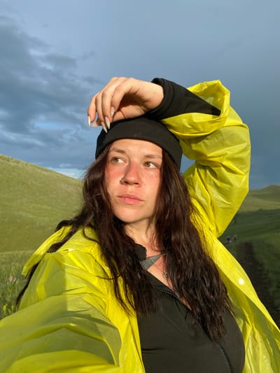 A woman, likely in her early 20s, is captured in a close-up, self-portrait style photograph against a backdrop of rolling green hills and a dramatic, cloudy sky. She is wearing a black beanie and a bright yellow raincoat. Her left arm is raised, with her hand resting on her forehead, and she is looking off to the right of the frame. Her long, dark, wavy hair frames her face, which features a nose ring and freckles. The lighting suggests it's either early morning or late afternoon, with a golden hue illuminating the scene. In the background, a dirt path winds up one of the hills, and two figures can be seen walking along it. The location is identified as Верхнеозёрное, Russia.