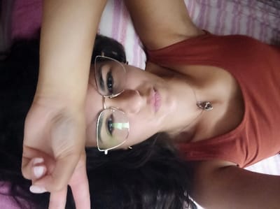 The scene depicts an adult female, lying supine, facing the camera, within an interior location in San Marzano sul Sarno, Italy. She has dark hair and a medium complexion. She is dressed in a reddish-brown tank top and wears clear-framed eyeglasses with a metallic bridge, through which her eyes are visible, with a green reflection present in the viewer's left lens. Her lips are light pink. A silver-toned necklace featuring two interlocked heart pendants rests on her clavicle area. Her left hand is elevated, positioned near her head, forming a 'V' gesture with the index and middle fingers, displaying light-colored nail polish. The immediate background consists of a striped textile, exhibiting pink and white hues, consistent with bedding or upholstery.