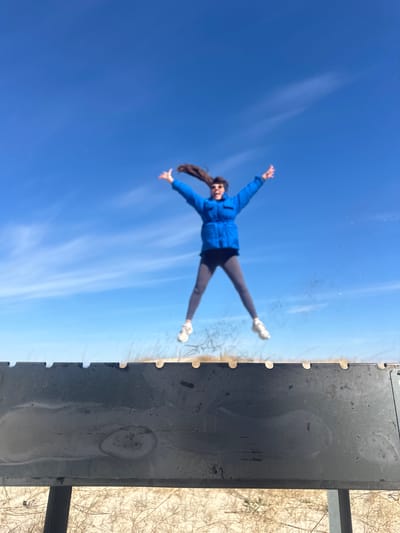 A woman wearing a blue jacket and grey pants is lying horizontally on a glass surface, arms and legs outstretched. Above her is a blue sky with white clouds. To the right of the glass surface is a dark grey structure, possibly metal. Beyond the metal structure is a snowy or sandy ground with bare tree branches. The location is likely a high viewpoint, possibly a rooftop or a viewing platform, in Zīvartiņš, Latvia. The woman's posture gives the impression of floating in the sky.