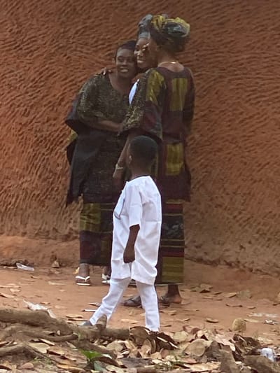 The image depicts a scene in Ochadam, Nigeria, featuring four individuals against a reddish-brown, heavily textured wall. In the middle ground, three adult females with dark skin tones stand in close proximity. The leftmost female, wearing a dark patterned top and wrapper with light open-toed sandals, smiles while embracing the central female with her left arm. The central female, dressed in a dark patterned outfit and matching headwrap, smiles and is embraced by both other women. A light-colored wristband is visible on her left wrist. The rightmost female, seen partially from the back, wears a similar dark patterned ensemble with a headwrap and dark sandals; her right arm is around the central female, and a subtle necklace is visible. All three women display expressions indicative of amusement or happiness. In the foreground, a young male child, also with dark skin, stands with his back to the viewer, observing the women. He is clad in a white traditional tunic and trousers. The ground consists of reddish-brown soil, sparsely covered with dry leaves, small branches, and roots, with a white plastic wrapper visible on the left.