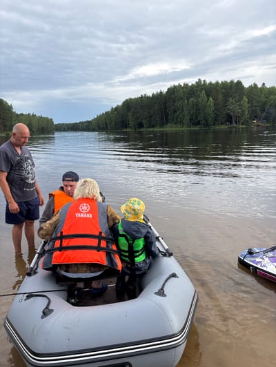 An overcast day scene depicts four individuals near an inflatable boat on the shore of a body of water, likely a lake or river, with a dense forest lining the opposite bank. The location is identified as Anteplevo (Okhotinskoe S/P), Russia.

In the foreground, a grey inflatable boat rests partially submerged in shallow, murky water. A fishing rod is placed across the boat's interior. To the left, a bald man wearing a dark t-shirt and blue shorts stands on the shore, facing the boat.

Inside the boat, three people are visible. A man wearing an orange life jacket and a black baseball cap is seated in the middle. To his right, a person with blonde hair also wears an orange life jacket with "YAMAHA" branding. In front of them, a child in a green life jacket and a yellow patterned hat is seated.

The water has a rippled surface, reflecting the cloudy sky. The opposite bank is densely covered with green trees. Further to the right in the water, a paddleboard with colorful graphics is visible. The overall atmosphere suggests a casual outdoor activity, possibly fishing or preparing for a boat ride.
