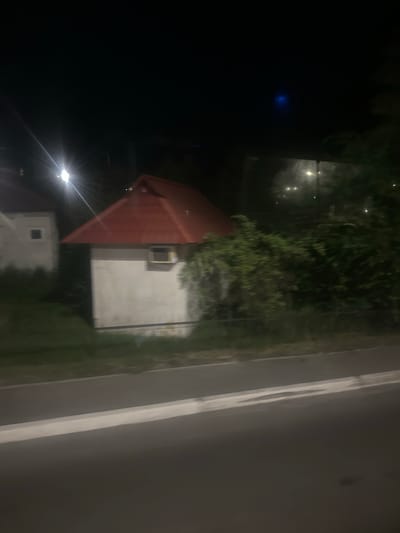 The image shows a nighttime view of a small, white building with a red, pitched roof. The building has a single, small, square window with what appears to be an air conditioning unit installed. To the left, another similar building is partially visible, also with a square window. Both structures are situated on a grassy area adjacent to a paved road. The road has a distinct white line marking its edge. Behind the buildings, lush green foliage and trees are visible, interspersed with a few blurred lights that suggest other structures or streetlights in the distance. The overall impression is of a rural or semi-urban setting at night.