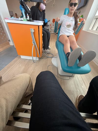 In a brightly lit dental office, a young girl wearing sunglasses sits in a teal dental chair, her legs crossed. She is wearing a white t-shirt with a cartoon cat graphic and black shorts. A dental professional in dark scrubs and a light pink mask stands behind her, wearing blue gloves and holding medical equipment. The office features a prominent orange dental cart with attached tubing. In the foreground, the legs of two individuals are visible, one wearing tan corduroy pants and the other in dark denim jeans. The floor is made of light-colored wood laminate. The overall impression is a sterile yet modern dental environment.