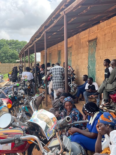 In Kassa, Nigeria, a group of people are gathered outdoors under a covered structure with a corrugated metal roof supported by wooden beams and metal poles. The building behind them is constructed of rough-hewn, tan bricks with green, slatted doors. A row of motorcycles, some with chrome detailing, are parked in front of the group, with one prominent motorcycle in the foreground displaying a poster. The poster features a portrait of a man named GWOM JERRY JOHN and text indicating he is deceased. People are dressed in a variety of clothing, including traditional attire and casual wear. Some individuals are seated on the ground, while others are standing or leaning against the motorcycles. The atmosphere appears somber, with some individuals looking thoughtful or sad. The environment suggests a public gathering, possibly a wake or funeral service, given the memorial poster. Trees and foliage are visible in the background, and the sky is overcast.