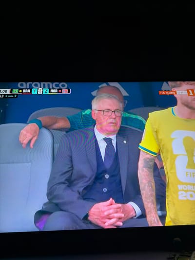 The image shows a screenshot from a soccer match broadcast, displayed on a television. In the foreground, a man in a dark suit and tie with glasses is seated, looking slightly to his left. His hands are clasped in his lap. To his right, another person, partially visible and wearing a yellow shirt, is also seated. Behind these individuals, a person in a green shirt with a blue wristband is leaning on a seatback, their arm extended. A scoreboard graphic at the bottom of the screen shows "0:00", flags of Brazil and Norway, and the score "BRA 1 2 NOR". Another graphic at the top right indicates "НА ЖИВО Б.Н.Т. 1". The background consists of blue seating and a dark ceiling. The image is not from the city of Ognianovo, Bulgaria; it is a televised sporting event.