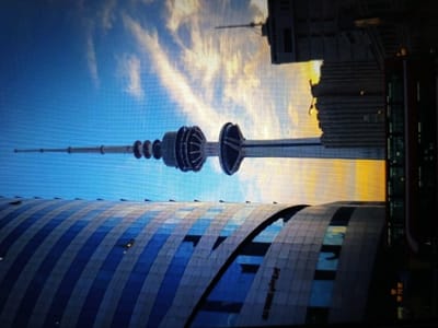 The image shows a view of a city skyline. In the foreground, a curved, modern building with blue and white striped panels is visible. Behind this building, a tall, slender tower with multiple levels and a distinctive spherical structure near its top dominates the center of the frame. The tower is set against a bright blue sky with wispy clouds. In the background, several high-rise buildings are visible, some appearing older and more industrial, while others are modern skyscrapers. The overall lighting suggests it is either sunrise or sunset, with warm orange and yellow hues in the sky. The image is captured from a slightly elevated perspective.