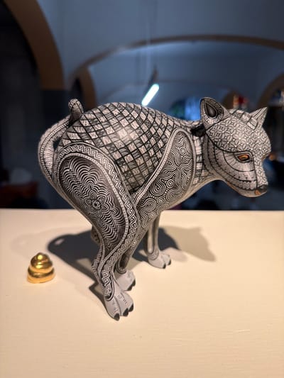 An Alebrije wolf figure, meticulously decorated with geometric patterns in black and white, sits on a pale wall, casting a shadow. The figure's head is tilted upwards, and its orange eyes stand out against the monochromatic design. A golden door knob is visible below, contrasting with the figure's intricate detailing and adding a touch of color. The background is blurred, suggesting an indoor setting.