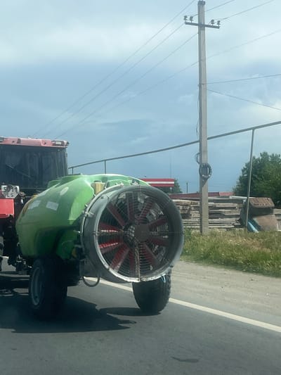 The image, rotated to its side, depicts a large green agricultural sprayer, likely an air blast sprayer or mist blower, mounted on the back of a red vehicle, possibly a truck or trailer. The sprayer features a prominent fan with red blades enclosed in a metal guard at its rear, and large black tires.

The scene is set outdoors on what appears to be a paved road with a white solid line marking the edge. To the right of the road (as viewed in the sideways orientation), there is a strip of grassy land. Overhead, multiple utility poles with power lines stretch across the sky, indicating an open, possibly rural or semi-rural, environment. In the background, obscured by the sprayer and utility pole, are trees and some structures, possibly industrial or storage, with stacked materials.

The weather is bright and clear, suggesting daytime, with some light clouds visible in the blue sky. Strong shadows cast on the road by the sprayer and vehicle indicate bright sunlight.

A notable detail is the text "5011 LS" visible on a red plate attached to the rear of the sprayer, resembling a license plate or identification tag. No specific activity is occurring; the vehicle and sprayer appear to be stationary. The location is Ayntap, Armenia.