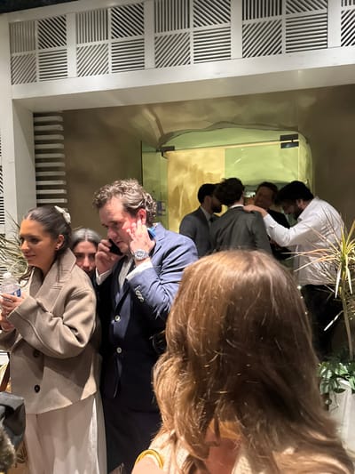 A group of people are gathered indoors. A man in a navy blue suit is talking on a cell phone, his wrist adorned with a watch. A woman in a tan coat stands beside him, holding a water bottle. Reflections of other people can be seen on a mirrored surface, including a man in a white shirt. A woman with brown hair is prominent in the foreground. The setting appears to be an upscale event or gathering.