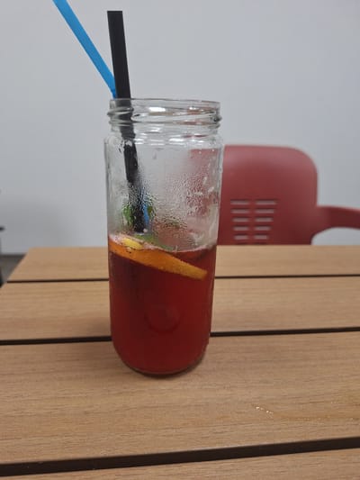 A refreshing, cold beverage served in a clear glass mason jar is the central subject of the image. The drink is a deep red color, likely a fruit punch or sangria, and contains slices of orange or other citrus fruit, along with subtle hints of green leaves, possibly mint. The glass jar is visibly covered in condensation, indicating the beverage is chilled.
Two straws are inserted into the drink: one standard black straw and one bright blue straw. The jar rests on a light-colored wooden surface, which appears to be composed of vertical planks, suggesting either a table or wall paneling.
In the blurred background, a prominent red object with a distinct curved shape and a pattern of vertical slits is visible. This object is set against a plain light gray or white wall. The scene suggests a casual setting, possibly indoors at a cafe, restaurant, or home. There are no people visible, and no text is present in the image. The image itself is originally oriented horizontally, rotated 90 degrees clockwise. The implied location context is Kraishte, Bulgaria.