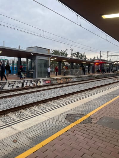 This image depicts a train station platform and tracks, viewed from an elevated position or an adjacent platform, looking across the railway lines. The setting is a public transport hub in Sant Adrià De Besòs, Spain.
**Subjects:** The foreground is dominated by several train tracks set into gravel, with intricate overhead electrical catenary wires crisscrossing against a muted, overcast sky. On the immediate right, a section of a platform is visible, featuring a textured grey walking surface, a distinct yellow safety line, and reddish brick paving further back. A round, metal manhole cover is embedded in this platform. On the opposite side of the tracks, another platform is populated by numerous individuals, mostly adults, either standing or seated beneath a modern shelter with glass panels. Station infrastructure like supporting pillars and what appears to be a fence are also present. Trees and distant structures punctuate the background beyond this platform.
**Activity or event:** The scene portrays typical daily activity at a train station, with people waiting patiently on the platform, likely for an arriving train.
**Notable details:** The sky is overcast, suggesting a cloudy day, possibly early morning or late afternoon. A prominent red signal light is visible on the far platform. The concrete barrier or wall adjacent to the tracks on the far side is marked with extensive graffiti.
**Visible text:** A small red sign or sticker positioned near the tracks on the far platform clearly reads "P. STOP". Indistinct graffiti lettering is also visible on the wall beside the tracks.