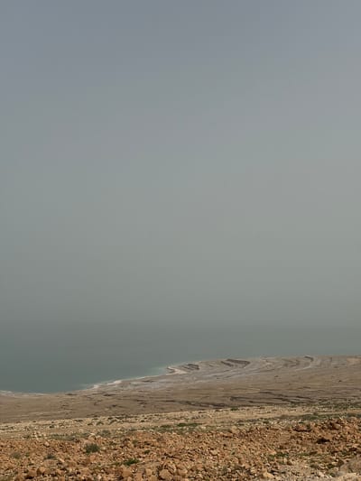 The image shows a landscape with a body of water on the right, partially obscured by land formations. The land is rocky and barren with some sparse vegetation. The body of water is calm and gray. The sky is overcast. The location is the city of Megilot Yam HaMelakh Regional Council, Palestinian Territory.