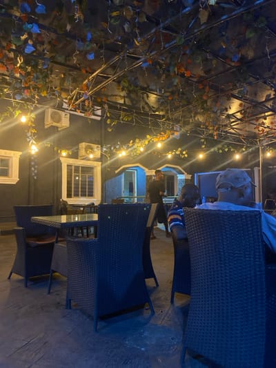 The image depicts an outdoor dining or lounge area at night in Igbessa, Nigeria. An overhead metal trellis structure is densely covered with artificial foliage in various shades of green, orange, and blue, interwoven with numerous string lights featuring exposed incandescent bulbs.
The background consists of a dark-colored building facade with several white-framed windows, one equipped with horizontal blinds. Two white wall-mounted air conditioning units are visible on the building. To the right, a doorway with a light-blue frame is partially visible.
In the mid-foreground, a square table with a glass top and a dark woven base is surrounded by two matching dark woven chairs, one with a light-brown cushion. To the right, two men are seated. The man in the immediate foreground is seen from behind, wearing a light-colored, possibly camouflage, cap and a light-blue shirt. To his left, another man is seated, partially visible, wearing a blue, orange, and white striped polo shirt, with his head tilted downwards. Further back, near the blue-framed doorway, a third man in a dark short-sleeved shirt and dark trousers stands facing away from the camera.
The ground is a dark, textured surface, possibly concrete or paving. The scene is illuminated primarily by the overhead string lights, casting a warm glow and creating a dim, evening ambiance.