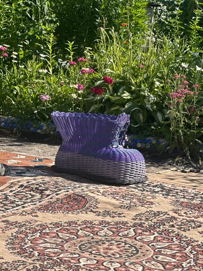 A woven purple basket sits on a rug with a detailed pattern. The rug is laid out on a textured surface, possibly stone or concrete, near a lush garden. The garden is filled with vibrant green foliage and various flowering plants, some with pink and white blossoms. The sunlight suggests it is daytime, and the bright light indicates fair weather. The overall scene appears to be outdoors, in a garden or patio setting, possibly in a residential area given the presence of plants and a rug. There are no people visible in the image. The basket has a metallic handle. A patch of blue and white mosaic tiles is visible near the base of the basket, peeking out from beneath the plants. The rug has a complex, traditional design with intricate motifs in shades of brown, red, black, and cream.