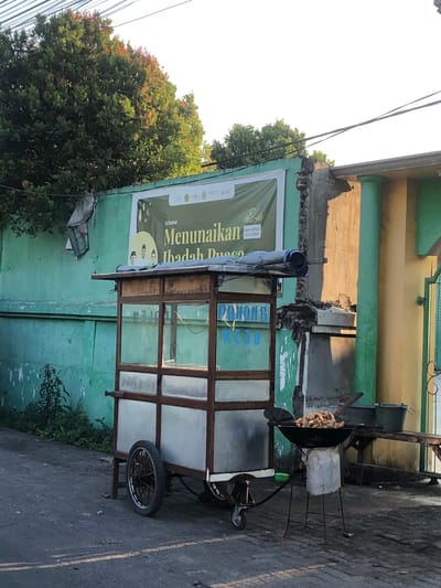 The image depicts an outdoor scene, likely in the city of Kalidawir, Indonesia, featuring a food cart and cooking equipment. In the foreground, a portable food cart constructed with a wooden frame and glass panels is visible, with one large wheel and a smaller front wheel. Next to the cart, a black wok rests on a tripod stand, filled with what appears to be fried food. A wooden bench and two buckets are positioned behind the cooking area. A green wall serves as a backdrop, adorned with a banner that reads "Selamat Menunaikan Ibadah Puasa." Overhanging the scene are tree branches with red-tinged leaves. The ground is paved with asphalt and some bricks. The overall atmosphere suggests a street-side food vendor preparing to serve customers.