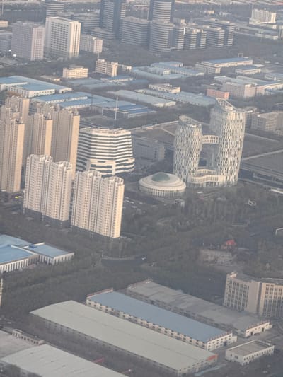 The image shows an aerial view of a dense urban landscape, likely a part of the city of Ürümqi, China. The dominant feature is the presence of numerous high-rise buildings arranged in a grid-like pattern. These buildings vary in design, with some exhibiting a repetitive, modular structure and others displaying unique architectural features like rounded tops and decorative facades. A central feature is a cluster of buildings with a distinct architectural style, including a cylindrical building topped with a dome. The streets are lined with trees, creating a dense green buffer between the buildings. The overall scene conveys a sense of high urban density and a modern urban environment.