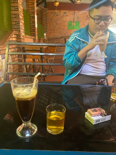 The scene depicts a young adult male seated at a black, reflective table within a cafe in the city of Đak Đoa, Vietnam. The male, wearing glasses and a teal athletic jacket over a white t-shirt, is actively drinking a light-colored beverage through a straw from a tall glass held in his left hand, while holding a smartphone in his right. On the table in the immediate foreground, from left to right, are a tall glass containing a dark, foamy beverage with a white straw; a small shot glass filled with a clear yellow liquid; and a white pack of "EAGLE" brand cigarettes with a pink and orange lighter resting on top. The background reveals exposed reddish-brown brick walls, partially covered by sections of artificial green turf, and black metal railings. Beyond the railings, additional wooden tables and chairs are visible. An orange paper lantern hangs from the ceiling.