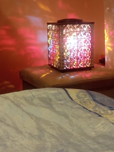 An indoor scene is dominated by a decorative, square lantern-style light fixture positioned centrally on a padded, light brown surface. The lantern features four sides composed of intricate, repeating geometric cutout patterns, allowing multi-colored light (primarily white, pink, blue, and yellow) to emanate brightly from within. This light casts distinct, fragmented, and colorful patterns onto the surrounding environment. The padded surface beneath the lantern, which appears to be upholstered furniture, displays smaller, scattered light projections. In the background, a solid-colored wall, primarily in warm orange and reddish hues, serves as a canvas for larger, more diffused, and undulating light patterns in shades of red, blue, and yellow. To the far right, a vertical, translucent object, possibly another decorative light source, is partially visible and also contributes to the light projections. The foreground is occupied by a textured fabric, light grey or bluish in color, possibly a bedsheet, featuring a patterned border in dark blue and yellow/gold, presented slightly out of focus. The image conveys a decorative, warmly lit indoor ambiance. The scene lacks specific visual identifiers or landmarks to confirm its location as Radishevo, Bulgaria. No individuals, specific actions, or direct interactions are depicted.