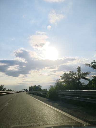 The image shows a view from a vehicle driving on a highway with a guardrail on the right and trees lining the side. The sky is bright with the sun peeking through some clouds. There are power lines visible across the sky. In the distance, there are a few cars and some signs. The road is asphalt with white lane markings. The overall impression is of a sunny day on a highway in or near Acerra, Italy.