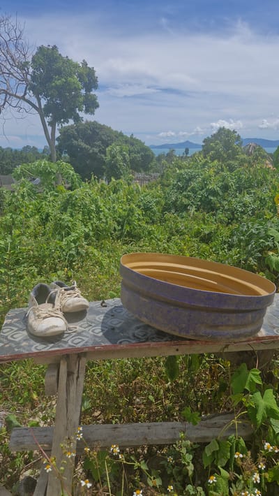 The image depicts a weathered wooden table outdoors in Ko Samui, Thailand. On the table are a pair of dirty white sneakers and a large, oval-shaped blue and yellow basin. The table itself appears makeshift, constructed from rough wooden planks. Behind the table, the scene is dominated by lush green vegetation, suggesting a tropical environment. In the background, trees and distant islands are visible under a partly cloudy blue sky. The overall impression is one of a rustic, outdoor setting.