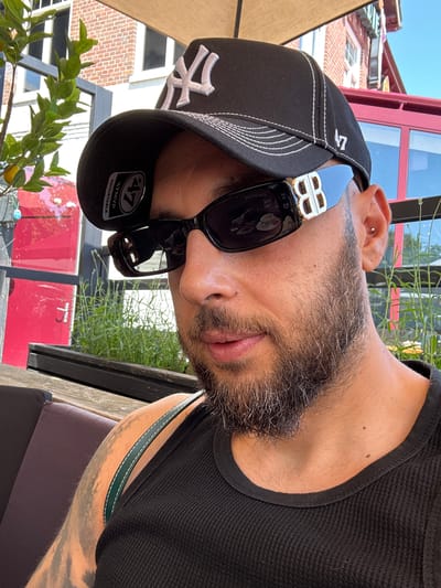 A man wearing a black New York Yankees baseball cap and black Balenciaga sunglasses is pictured in a close-up, waist-up shot. He has a beard and an ear piercing. The man is wearing a black ribbed tank top and has tattoos on his arm. He is seated at an outdoor table with a wooden bench behind him. In the background, there is a red building with windows, a small tree with lemons, and potted plants. The scene appears to be a cafe or restaurant setting. The image does not contain any information that would allow for identification of the specific location in Emmen, Netherlands.
