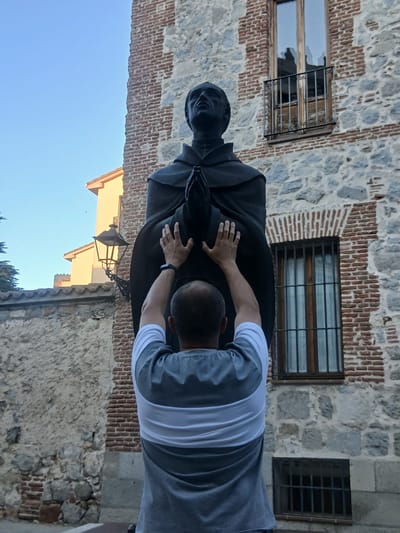 A man wearing a gray and white striped t-shirt is reaching up with both hands to touch the hands of a dark bronze statue of a robed figure with clasped hands. The statue is positioned outdoors in front of a weathered stone and red brick building. The building has multiple windows, one with a decorative black metal grate. To the left of the statue, a street lamp is mounted on the building. The sky above is clear and blue. The scene appears to be in the city of Ávila, Spain, likely near a religious or historical site given the statue and architecture.