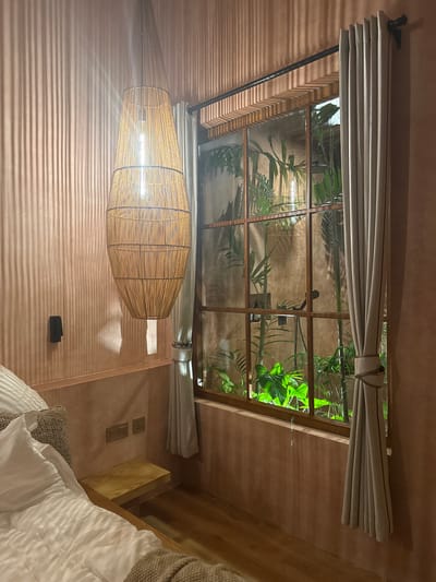 The interior of a bedroom in El Nido, Philippines is visible. A window with a grid-like frame dominates the upper part of the image, looking out onto lush greenery. A light grey curtain is drawn back on either side of the window. Below the window hangs a large, woven pendant lamp with a visible lightbulb. The walls are textured and painted in a warm, earthy tone. A bed with white linens and a textured throw is partially visible on the right side of the image. A wooden nightstand with electrical outlets is positioned next to the bed.