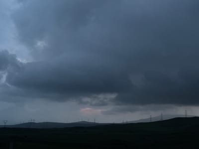 A wide-angle landscape photograph captures a dramatic, overcast sky over the hills surrounding Sisian, Armenia. The sky is filled with heavy, dark clouds, with a faint hint of pink light peeking through near the horizon. Silhouetted against the dim light are numerous tall, lattice-like power transmission towers dotting the undulating hills. The foreground consists of dark, uncultivated terrain, suggesting a rural or semi-rural environment. The overall mood is somber and brooding, indicative of impending weather. No people or animals are visible in the scene.