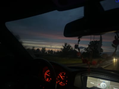 The image depicts a view from inside a vehicle driving on a road at dusk. The sky is a deep blue with streaks of pink and orange, indicating the sun is setting. Silhouetted trees line the side of the road. Inside the car, the dashboard displays glowing red instruments, and a central screen shows navigation information. The road ahead has its own headlights visible from other vehicles. The scene is peaceful and serene, capturing the transition from day to night.