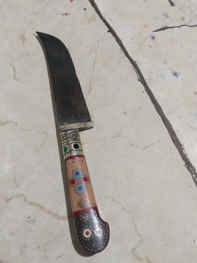 The image shows a curved, single-edged knife with an ornate handle lying on a marble-like surface. The blade is dark and appears metallic, with visible scratches. The handle is made of a light-colored material, possibly wood or bone, and is decorated with colorful circular patterns in red, blue, and black, as well as metallic accents. The surface it rests on is a light beige with brown veining and some darker speckles, suggesting it could be a countertop or tabletop. The lighting is somewhat dim, casting subtle shadows. There is a dark, twig-like object running diagonally across the upper right portion of the image. No people or specific location details beyond the mention of Shirin, Uzbekistan are visible.