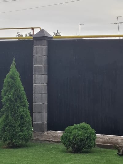 In the city of Kara-Balta, Kyrgyzstan, a fenced yard is captured under an overcast sky, suggesting rainy weather. A tall, conical evergreen tree stands to the left, adjacent to a stone pillar that supports a dark fence. To the right of the pillar, a low, rounded evergreen bush is situated on a manicured lawn. The fence itself is a dark, solid material, with vertical streaks indicating the presence of falling rain. A yellow pipe runs horizontally above the fence, supported by a rusty bracket attached to the stone pillar. In the distance, several television antennas are visible against the grey sky. The scene is quiet, with no people or animals immediately apparent, except for a partially visible patterned object in the lower right corner, which might be a decorative element or a small animal.