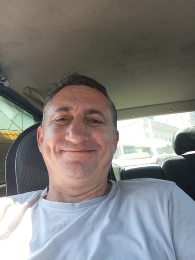 A medium shot selfie taken inside a car captures a Caucasian man in his late 40s to early 50s. He is seated in the driver's seat, facing the camera directly with a slight smile. The man has short, graying brown hair, a fair complexion, and prominent dimples on his cheeks. He is wearing a light blue t-shirt and a silver chain necklace. The interior of the car shows the headliner, a portion of the passenger seat with a patterned fabric, and the back seats. Through the rear window, an out-of-focus view of buildings and an overcast sky is visible. The scene is well-lit, suggesting it was taken outdoors during the daytime. The specific location within Vasylkiv, Ukraine, is not identifiable from the visual cues in the image.