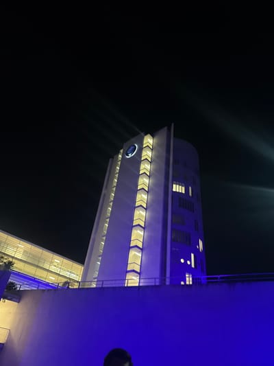 The image presents a nighttime scene in Ciudad López Mateos, Mexico, focused on a modern architectural structure. A tall, slender building stands prominently, featuring a circular, illuminated logo near its pointed apex. Below the logo, a vertical segment of the facade is adorned with a series of chevron-shaped, recessed windows or panels emitting a warm yellow-white light. An adjacent, curved section of the building displays various rectangular and circular windows, some of which are illuminated with similar yellow-white light. To the left, a multi-story structure with a glass facade, also internally illuminated, appears connected, possibly by a skywalk. In the immediate foreground, a broad concrete wall or barrier is strongly illuminated by blue ambient light. The very bottom center of the frame shows the top of an individual's head, out of focus. The upper portion of the image is dominated by a dark night sky, with distinct rays of light emanating from an unseen source in the upper right quadrant.