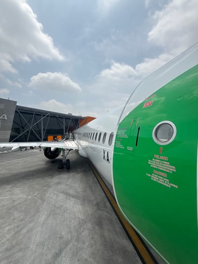 An airplane is prominently featured, parked at an airport gate, viewed from a low angle on the tarmac. The aircraft's fuselage is white with a distinctive bright green upper section and an orange tail fin, though only a small part of the tail is visible. A major focus is on an emergency exit door on the green section of the fuselage, which has a small porthole window.
The door features red text warnings and instructions in both Spanish and English. Visible text includes "SALIDA," "EXIT," and an arrow pointing upwards. Below this, there are instructions "PARA ABRIR" and "TO OPEN" with an illustration showing how to operate the door. Further right, "PELIGRO" and "DANGER" are printed, followed by a warning: "NO ABRIR LA PUERTA SI LA LUZ ROJA DE ADVERTENCIA SE ILUMINA INTERMITENTEMENTE (CABINA PRESURIZADA!)" and its English translation: "DO NOT OPEN DOOR IF RED WARNING LIGHT IS FLASHING (CABIN PRESSURIZED)." The registration "XA" is also visible on the white part of the fuselage.
The airplane's landing gear and part of an engine are visible, alongside a modern-looking terminal building with large glass panels and a connecting jet bridge structure. The ground is a concrete tarmac with yellow lines. The sky above is cloudy, suggesting a daytime scene. The location is Apodaca, Mexico. No people are visible.