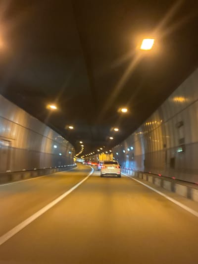 An interior shot taken from the perspective of a vehicle moving through a brightly lit tunnel. The tunnel has a dark, curved ceiling with rectangular lights spaced at intervals, casting a warm glow. The walls are textured and appear metallic or reflective, with vertical lines and a series of smaller lights embedded within them. The road is asphalt with a prominent white lane marker. Several vehicles are visible ahead, including a white car in the middle ground and others further in the distance. The overall impression is one of forward motion within a man-made subterranean passage. The city of Vysokoye, Russia is not visible or inferable from the image.