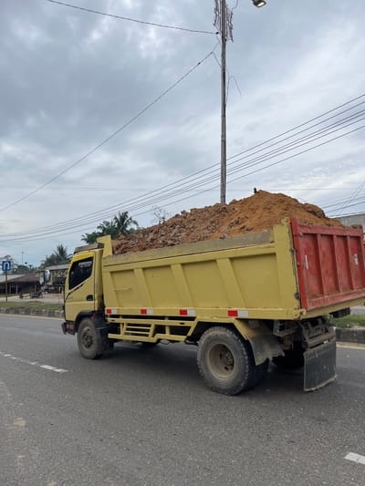 In the city of Tanahterban, Indonesia, a yellow dump truck is captured in motion on an asphalt road under an overcast sky. The truck is laden with a large pile of soil and rocks, which fills its bed and spills slightly over the sides. The truck's side is visible, showing red accents on the rear, and reflective safety strips on the yellow body. In the background, there are buildings, palm trees, a utility pole with wires, and a street sign indicating a U-turn is permitted. The overall scene suggests ongoing construction or land movement activities in the area.