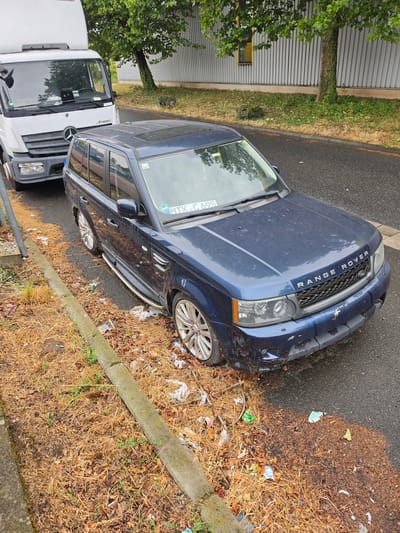 An eye-level, medium shot captures a dark blue Range Rover Sport parked next to a white Mercedes-Benz Atego truck on a paved road in Ginsheim-Gustavsburg, Germany. The Range Rover is positioned in the foreground and appears to be slightly angled towards the viewer. The front passenger side tire of the Range Rover is flat and appears to be deflated. The road is littered with debris, including dried leaves, grass, and various pieces of trash. A concrete curb runs along the side of the road, separating it from a strip of dry, brown grass and more scattered debris. Behind the Range Rover, the white truck is parked, facing away from the viewer and slightly to the left. The truck's front grille, headlights, and windshield are visible. In the background, a mature tree with green foliage stands tall, with a grey metal building facade extending behind it. The overall scene suggests an urban or industrial area, with the discarded waste indicating a lack of proper maintenance or sanitation.