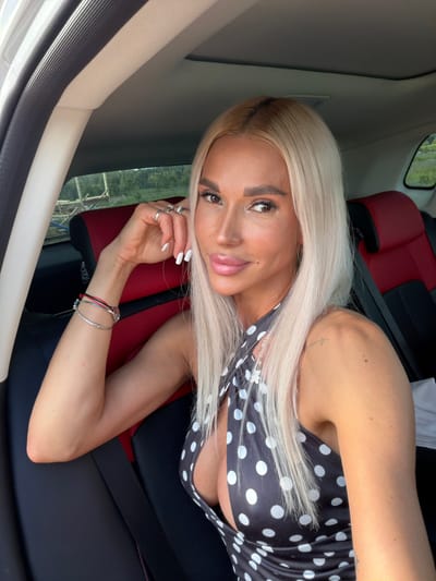 A young woman with long, straight blonde hair and a light complexion is captured in a close-up, waist-up portrait, seemingly taken inside a car. She is positioned leaning against a dark and red car seat, looking directly at the camera with a slight, alluring smile. Her eyes are dark brown and lined with prominent eyeliner and long lashes, complemented by bold, well-groomed eyebrows. Her lips are full and glossy, with a slight upturn at the corners.
She is wearing a dark top adorned with white polka dots. Her left arm is bent, with her hand resting near her face, showcasing white-painted nails and multiple silver rings on her fingers. Her right arm is also bent, with several thin bracelets visible on her wrist, including silver bands and a red string. The interior of the car is visible around her, with a dark and red car seat and headrest in the background, along with a seatbelt. Through a window behind her, green foliage and what appears to be a fence or distant structure can be seen, suggesting an outdoor setting during daytime. The overall impression is a casual pose within a vehicle, with the woman as the central focus. No visible text is clearly legible in the image. The location context provided is Kon, Russia.